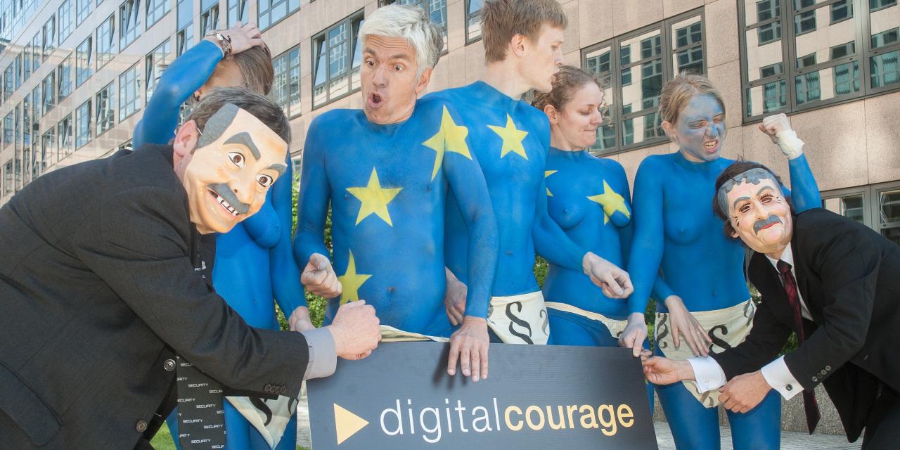 Mehrere nackt Menschen, die wie die Europa-Flagge angemalt sind (blau mit gelben Sternen). Daneben Menschen mit Masken. SIe halten ein Schild von Digitalcourage in den Händen.